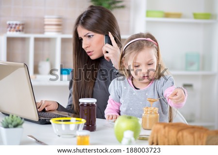 Overworked Business Woman and her little daughter in the morning. Mother read mail and make phone calls before going to work. Daughter smearing peanut butter on bread. - stock photo